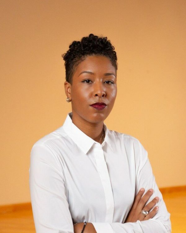A female-presenting person with dark skin poses in a white shirt against a light mustard colored background