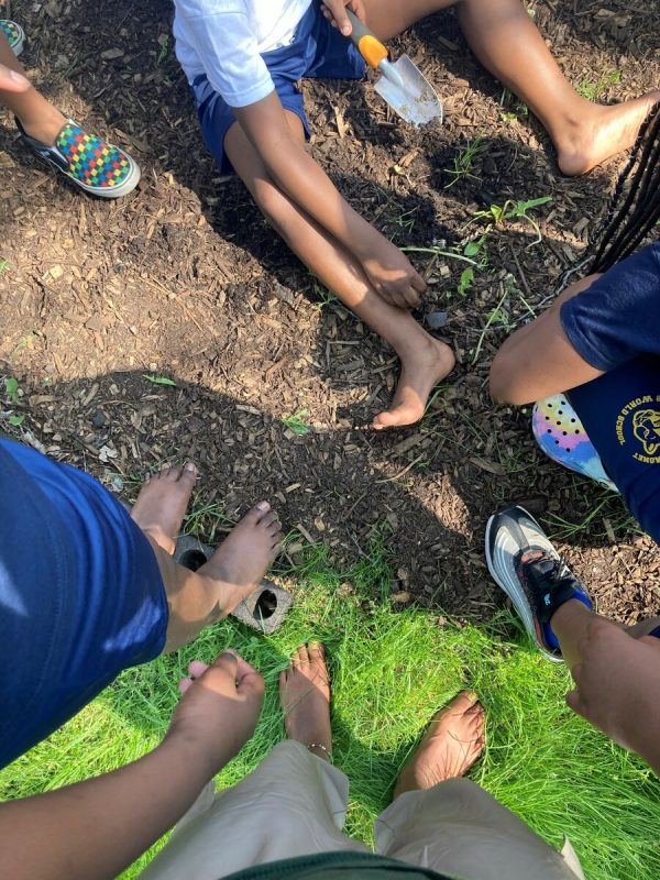 Several pairs of feet, some bare, some with shoes, on dirt and grass seen from above.