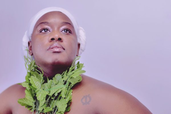 A person with medium-dark skin wearing a white headscarf and a necklace made of faux leafy greens raises their head and eyes upward, their eyes accented by shimmery makeup, against a lavender background. Though the image cuts off at the shoulders, their arms appear to extend outwards as if their hands are placed on their hips in a superhero pose.