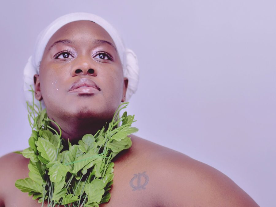 A person with medium-dark skin wearing a white headscarf and a necklace made of faux leafy greens raises their head and eyes upward, their eyes accented by shimmery makeup, against a lavender background. Though the image cuts off at the shoulders, their arms appear to extend outwards as if their hands are placed on their hips in a superhero pose.