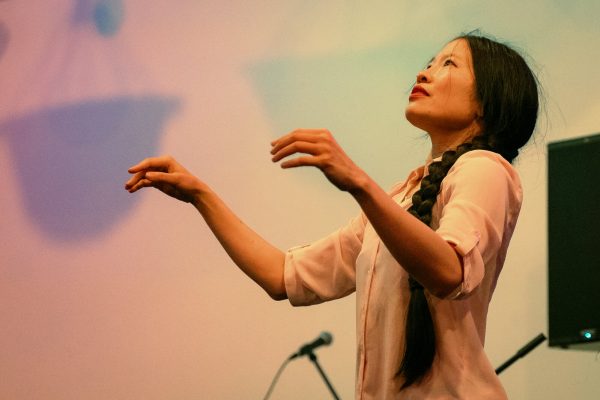 A female-presenting person with long dark hair styled in a braid that cascades over their left shoulder. They are turned to our left and look up, arms raised to shoulder level