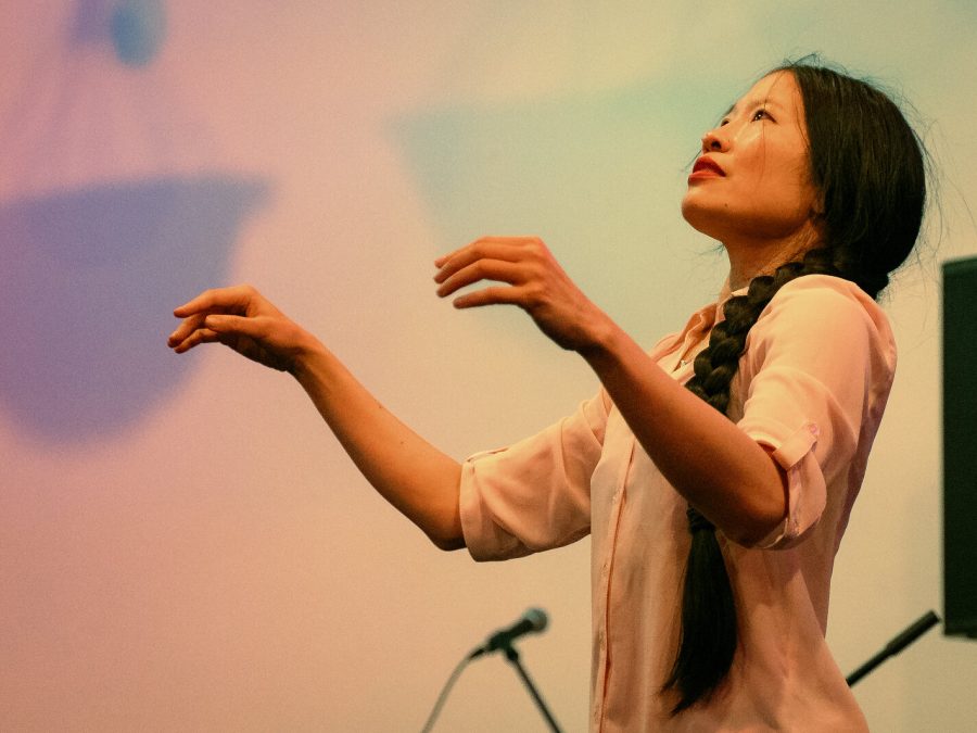 A female-presenting person with long dark hair styled in a braid that cascades over their left shoulder. They are turned to our left and look up, arms raised to shoulder level