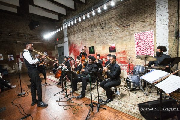 A saxophonist stands in front of a group of musicians playing a variety of other instruments in a brick-walled room