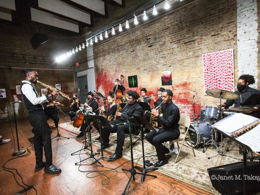 A saxophonist stands in front of a group of musicians playing a variety of other instruments in a brick-walled room