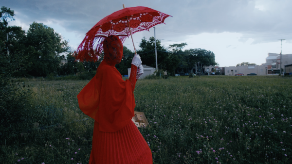 A person dressed in all red including their face covering holds a red lace parasol in a field on a cloudy day
