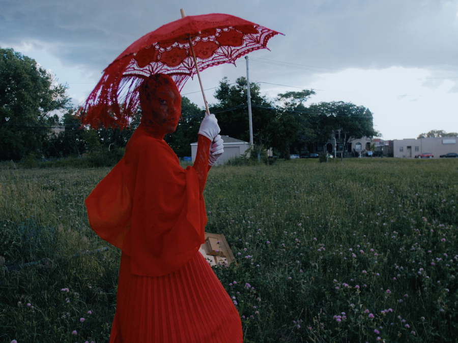 A person dressed in all red including their face covering holds a red lace parasol in a field on a cloudy day