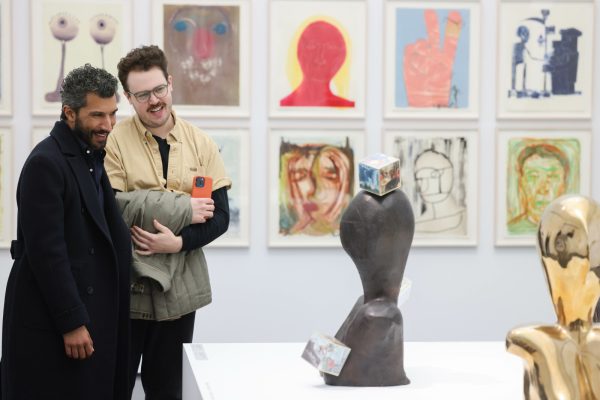 Two people look at a set of two sculptures in a gallery space with a series of drawings on the wall behind them.