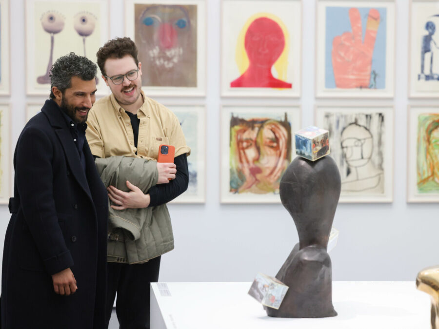 Two people look at a set of two sculptures in a gallery space with a series of drawings on the wall behind them.