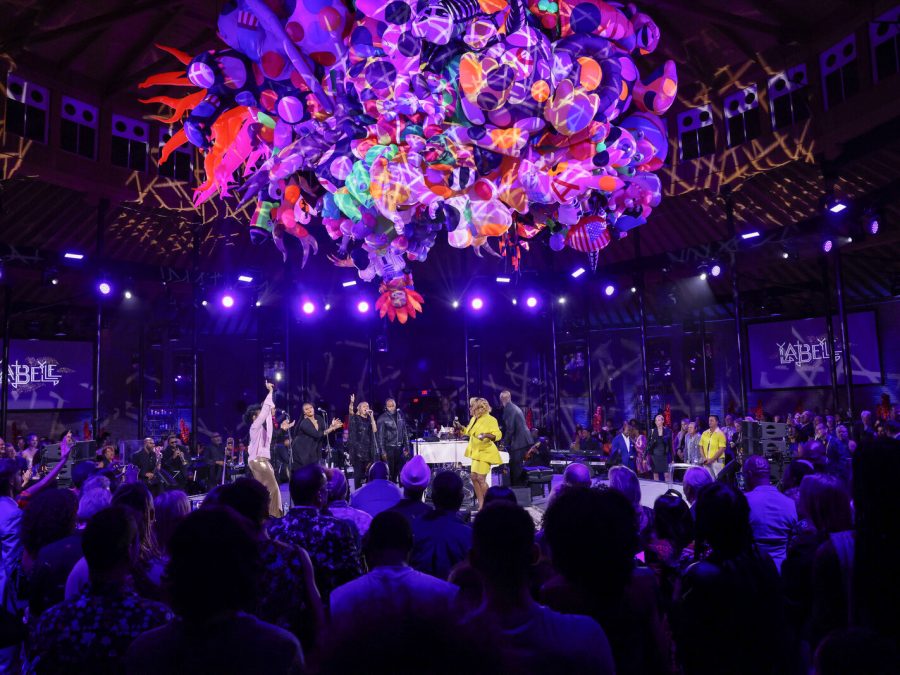 Photo of event with performers onstage with an art installation in purple, red, orange, green, and pink hanging above them.