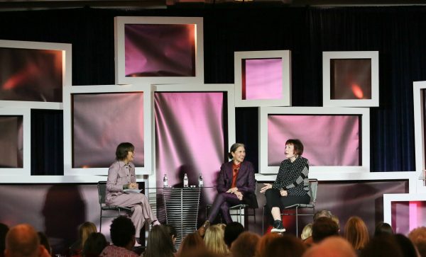 Three women sit on a stage surrounded by white-framed filled with pink fabric