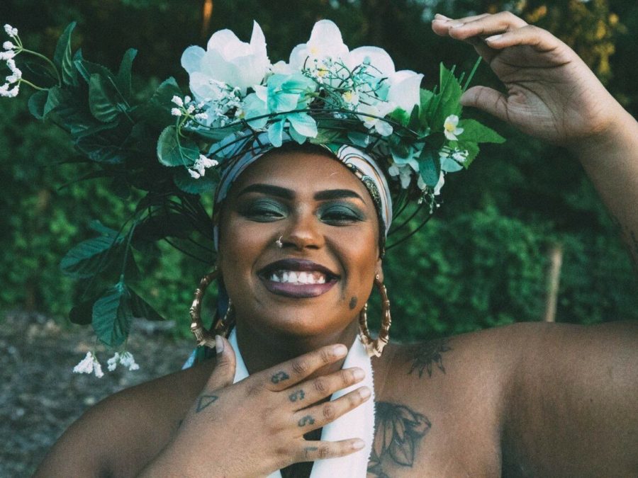 Close-up photo of a medium-skinned person wearing a floral headpiece. Their right hand is held below their chin with splayed fingers while their left hand is held in a c formation at the top of their head.