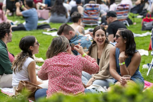 A smiling group of people sit on a blanket on a lawn enjoying each others company. They are surrounded by several other groups of people sitting on blankets.