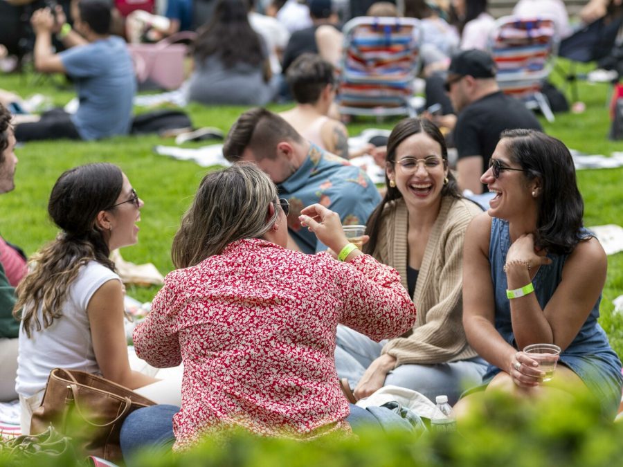 A smiling group of people sit on a blanket on a lawn enjoying each others company. They are surrounded by several other groups of people sitting on blankets.