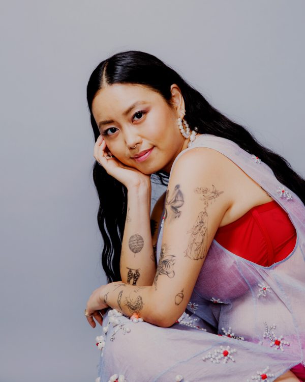 Photo of a person with long black hair parted in the center, with black tattoos on medium-light skin, wearing a red and lilac outfit.