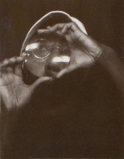 Greyscale image of a person wearing glasses and a hat holding their hands up to their face in an oval shape to frame it