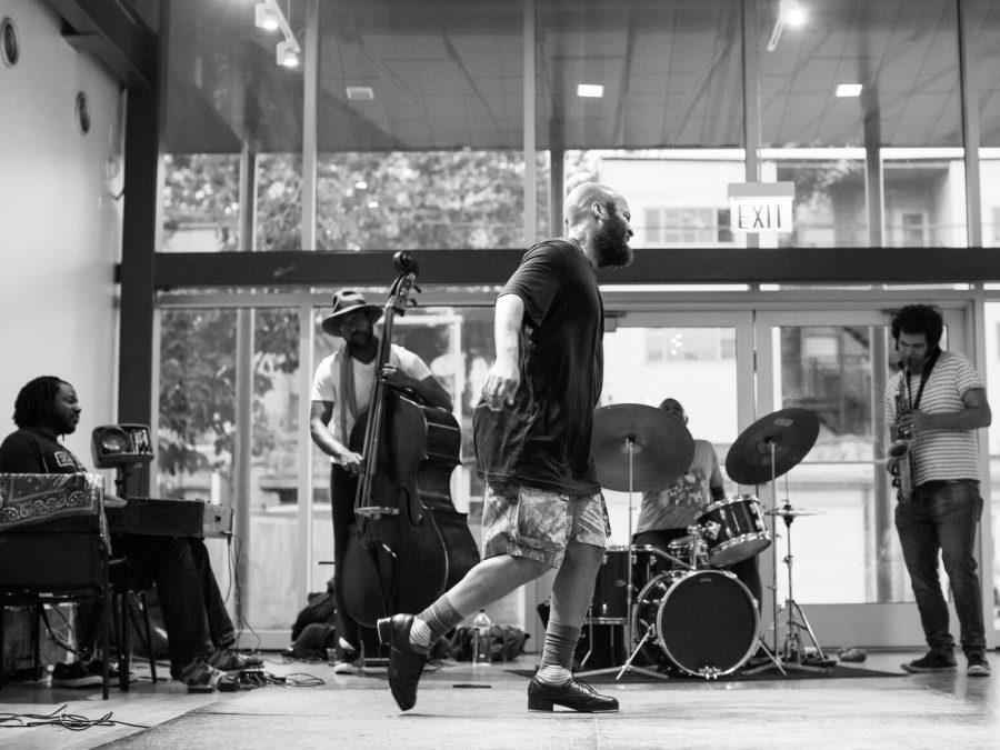 Black-and-white photo of a man tap dancing in front of five musicans.