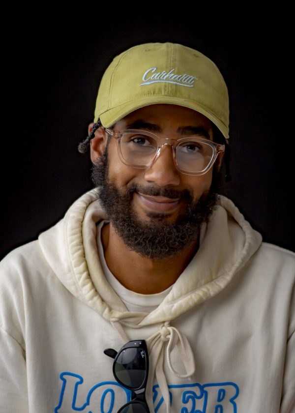 A male-presenting person with medium-dark skin wears a yellow hat, clear glasses, and a white hoodie with blue text reading Lover.