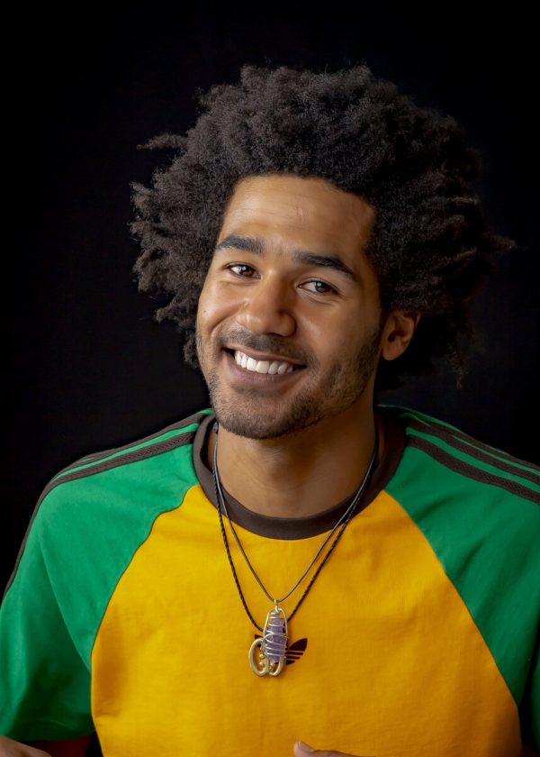 Portrait male-presenting person with medium-dark skin and a black afro who smiles at the viewer. They wear a yellow and green jersey and two necklaces.