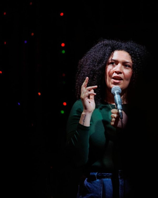 A dramatically lit woman stands in front of a microphone. She gestures with her right hand as she looks up and to her right, her mouth agape as if captured mid-sentence.