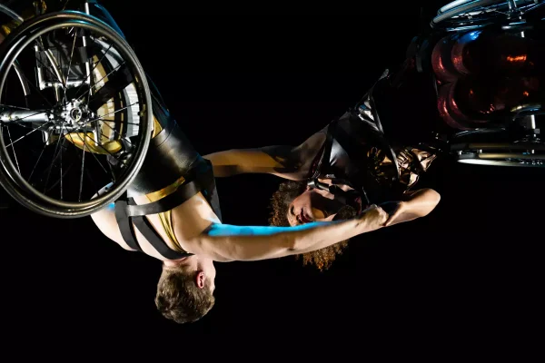 A close-up of Alice Sheppard and Laurel Lawson suspended in the air, arms outstretched and clasping each other's hands. Alice is a multiracial Black woman with coffee-colored skin and short curly hair; she wears a shimmery deep red costume. Laurel is a white woman with very short cropped hair; she wears a shimmery gold costume with thick black shoulder straps. The dancers are somehow upside down and horizontal at the same time, their wheels shining and facing out; if they let go, they will swing like pendulums.