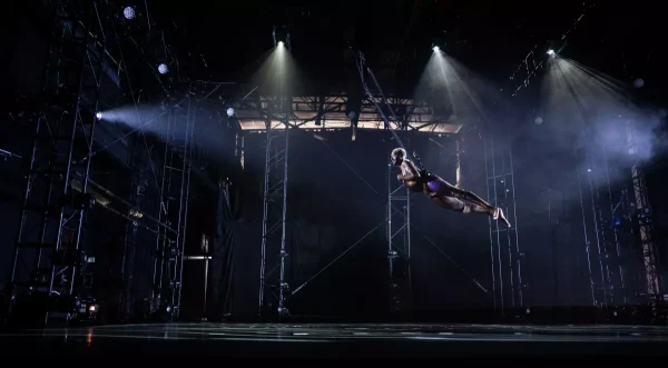 Jerron Herman soars high above the stage; launching across a high-ceilinged performance space. He is a dark-skinned Black man with blonde hair and a dark mustache; he wears a gold and purple shimmery costume as he flies, his long legs stretched behind him and arm tucked against his chest. White lights shine through the darkness; illuminating Jerron and the huge metal truss that surrounds him.