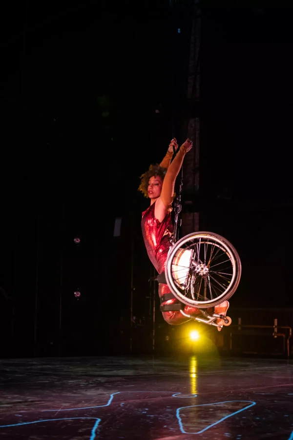 Alice Sheppard, a multiracial Black woman with coffee-colored skin and short curly hair, bounds through the air. Her deep red costume shimmers in the theatrical lighting, as a bright yellow beam appears under her. She arches her body, arms reaching up and hands gripping the cable that extends from her chair; her eyes are closed and her facial expression depicts a quiet exhilaration.