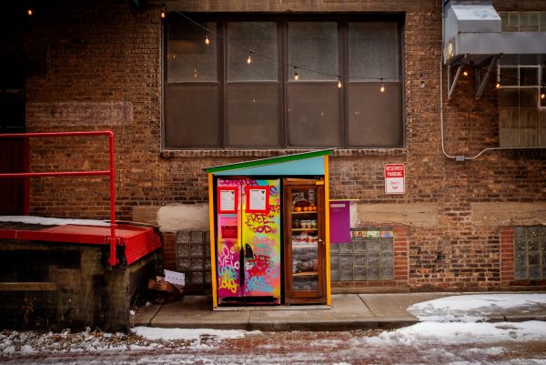 A colorful fridge outside a brick building with a red loading dock to the left. A small bit of snow appears on the ground.