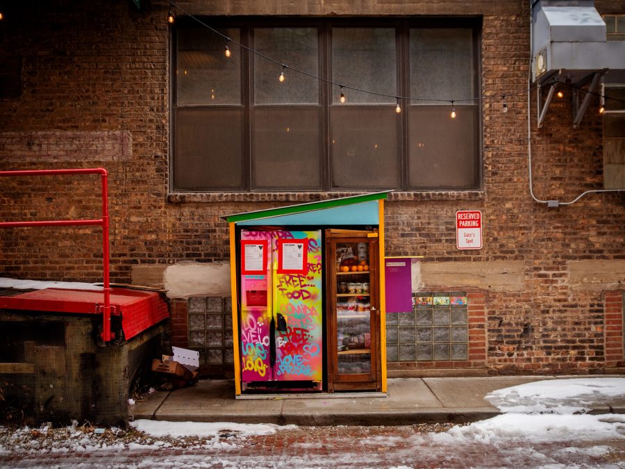 A colorful fridge outside a brick building with a red loading dock to the left. A small bit of snow appears on the ground.
