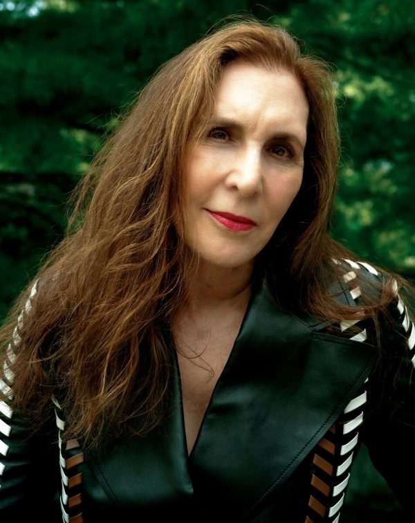 Close-up portrait of a woman with long brown hair gazing intently into the camera. She wears a green shirt against a green background.