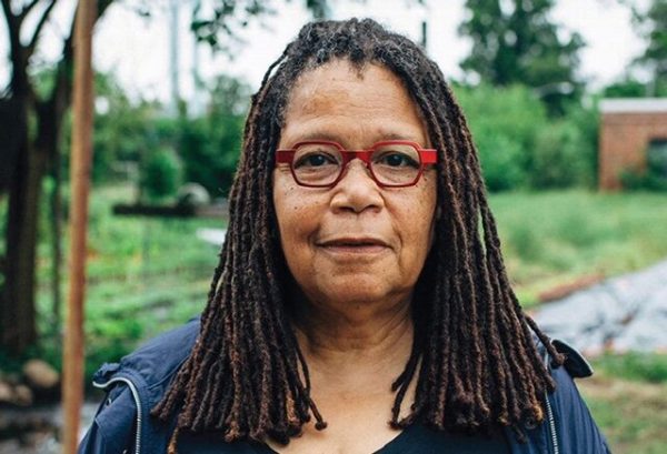 Three-quarter portrait of a female-presenting person with medium length brown dreadlocked hair and red glasses outside.