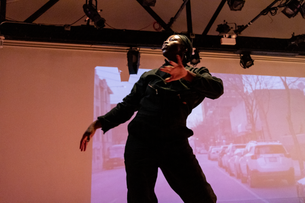 A female-presenting person with dark skin on a stage with a video projected behind them