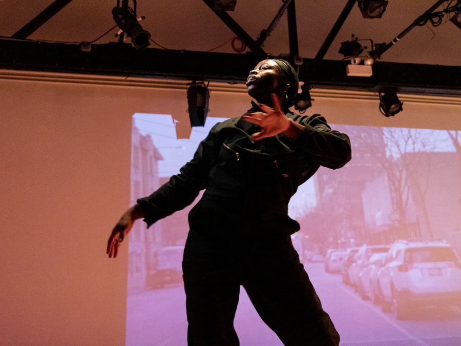 A female-presenting person with dark skin on a stage with a video projected behind them
