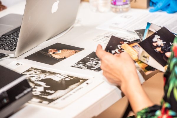A light skinned hand holds a photograph resting on a table filled with other photographs