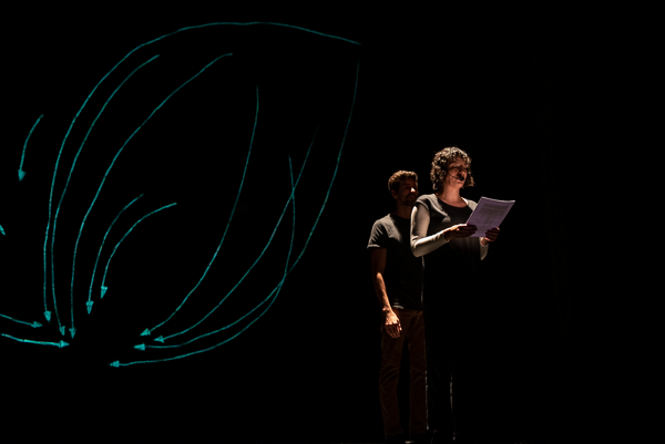 Two presenters dressed in black lit by theater lights standing in front of a dark backdrop with a variety of green arrows.