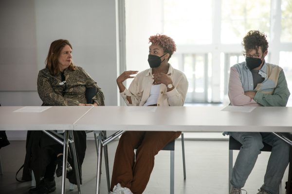 Three people sit on the same side of a long white table. The middle person seems to be engaged in conversation with the person on their right as they gesture with their hands.