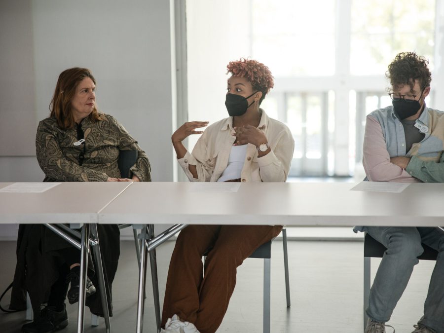 Three people sit on the same side of a long white table. The middle person seems to be engaged in conversation with the person on their right as they gesture with their hands.
