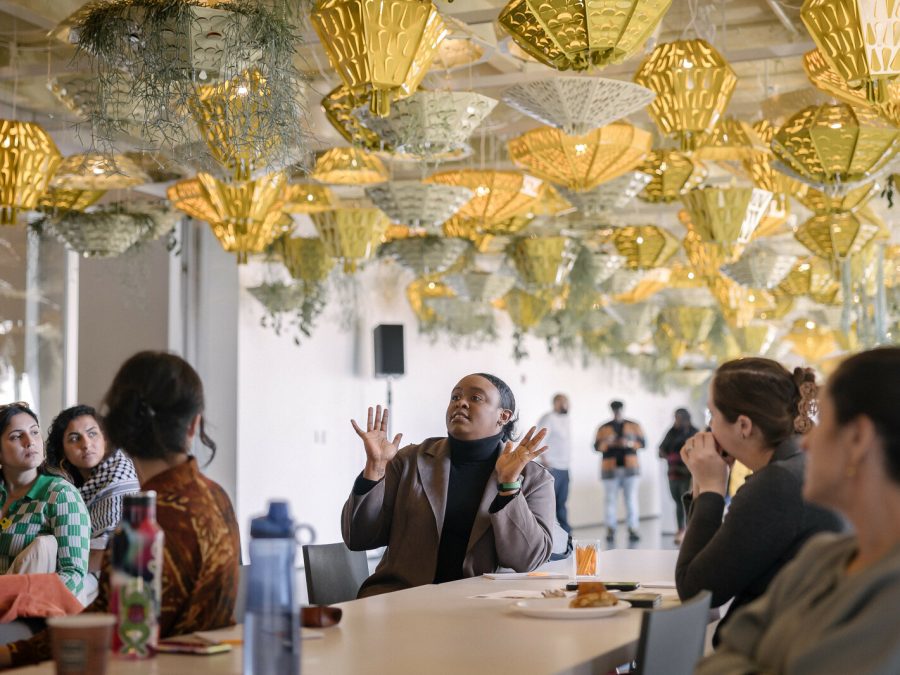 A group of people sitting at a table looking at one person who is gesturing with both hands as they seem to speak