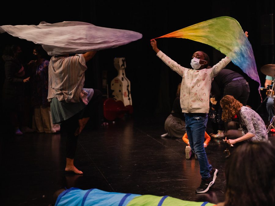 Two people raise large, colorful pieces of fabric over their heads on a darkened stage.