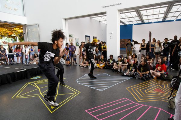 Two people energetically performing a dance move on a floor marked with colorful geometric shapes surrounded by an audience watching intently.