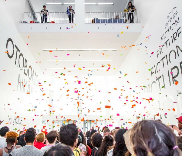 Colorful pieces of paper float over a crowd of people in a large, open white room.