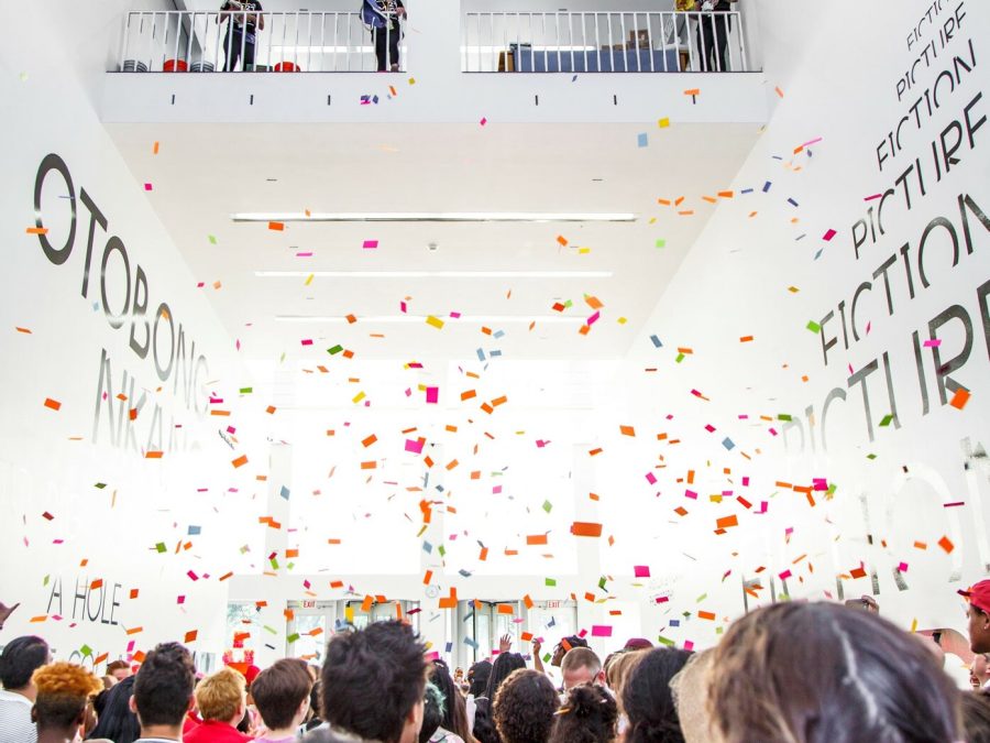 Colorful pieces of paper float over a crowd of people in a large, open white room.