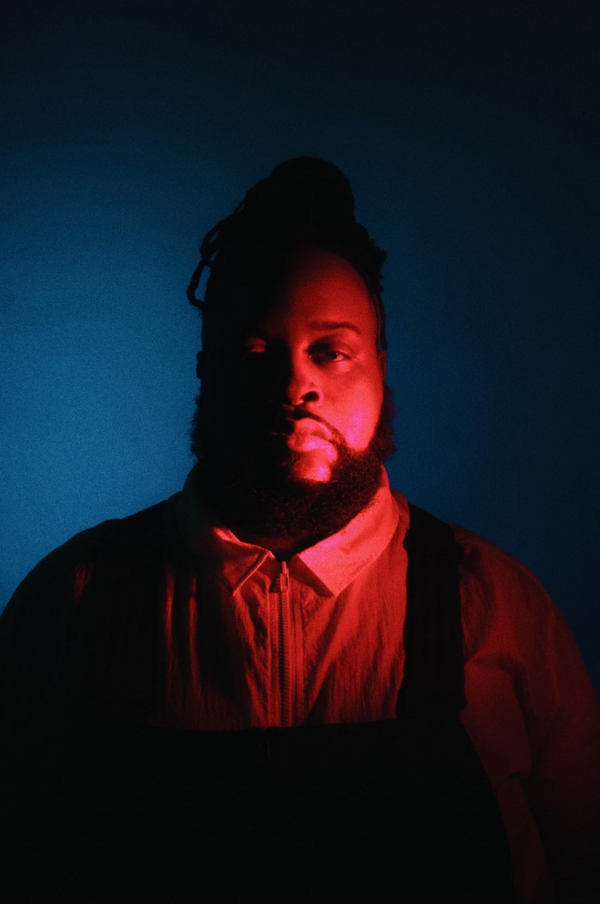 A three-quarter portrait of a well-built male-presenting person against a blue background who is dramatically lit from their left with a red light