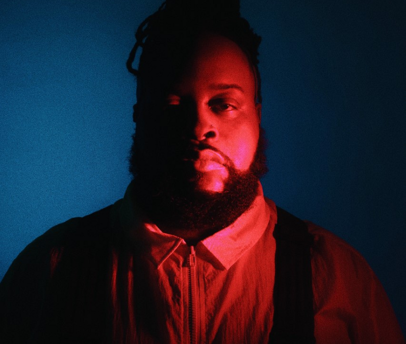 A three-quarter portrait of a well-built male-presenting person against a blue background who is dramatically lit from their left with a red light