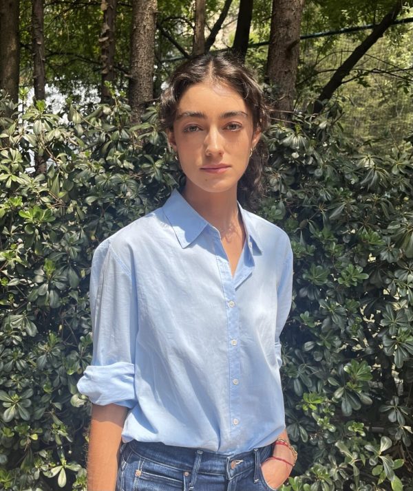 Portrait of a person in a light blue shirt with their left hand in their jean pocket in front of some shrubbery