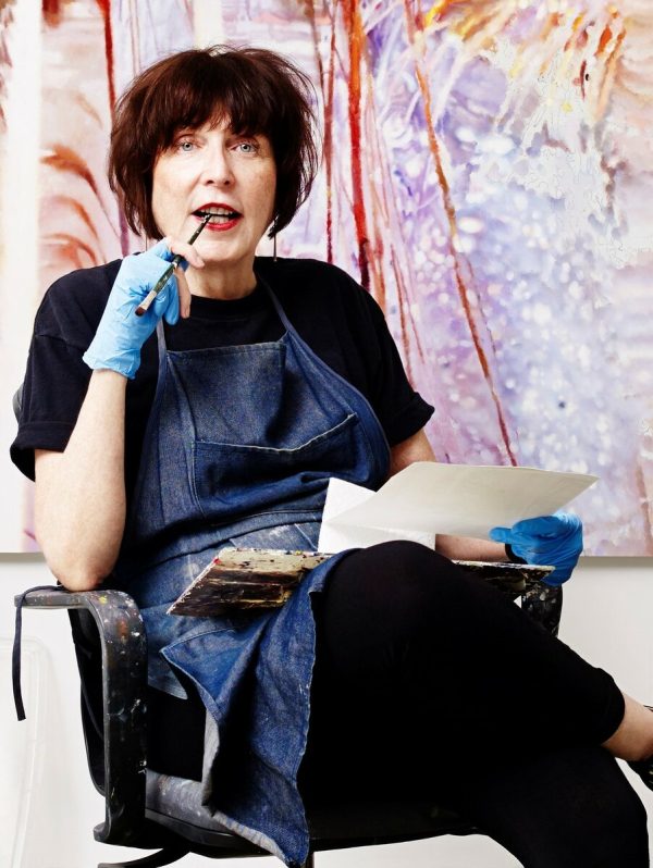 A woman with short brown hair sits holding a pencil in one hand and a piece of paper in the other.