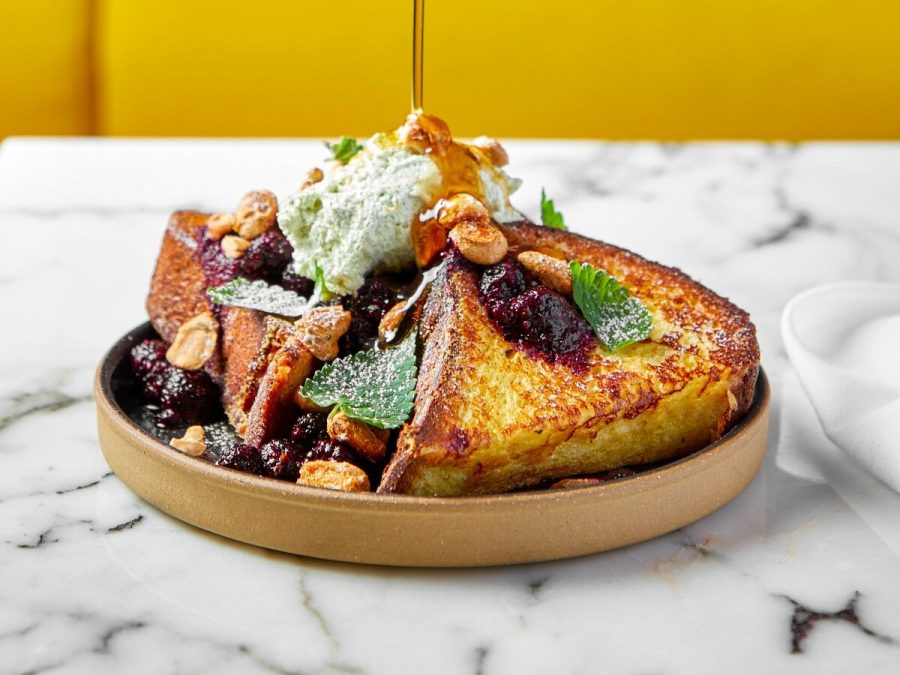 A stream of brown liquid is poured on top of a large serving of french toast with whipped cream and jam