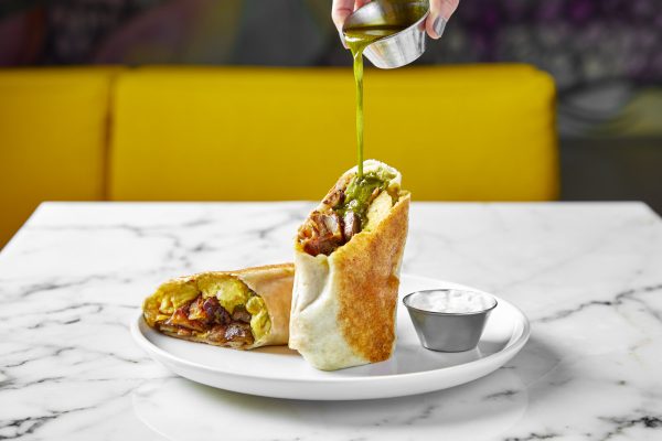 A burrito on a white place cut in half with one portion positioned upright and a sauce being poured into it. A side of sour cream is in a small metal dish next to it.