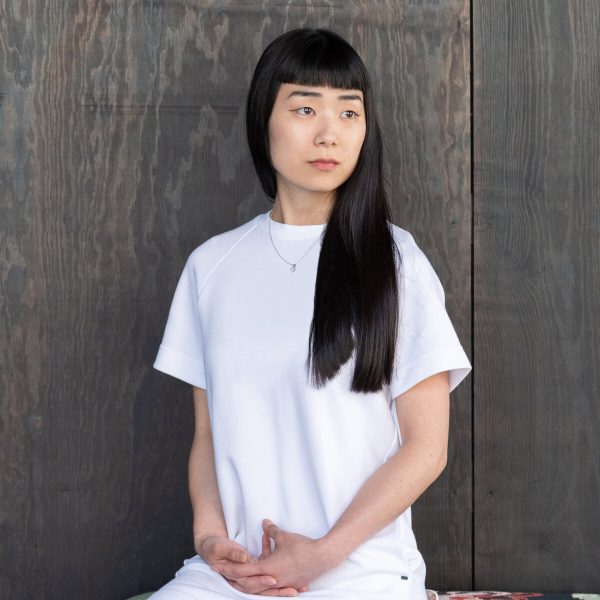 A female with long dark hair and square bangs poses against a wood plank wall. She is dressed in all white and looks off to her left.