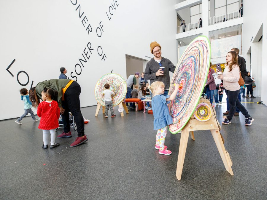 Kids and adults gather in an open, two-story space, many around easels will round canvases