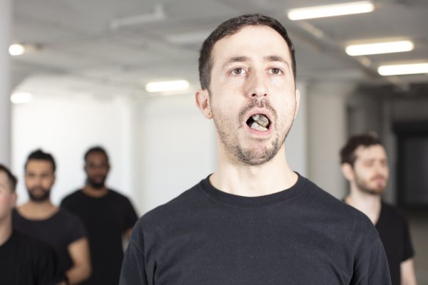 A group of people at varying distances from the camera are dressed in black shirts. The man in the foreground has his mouth open; inside are rocks.